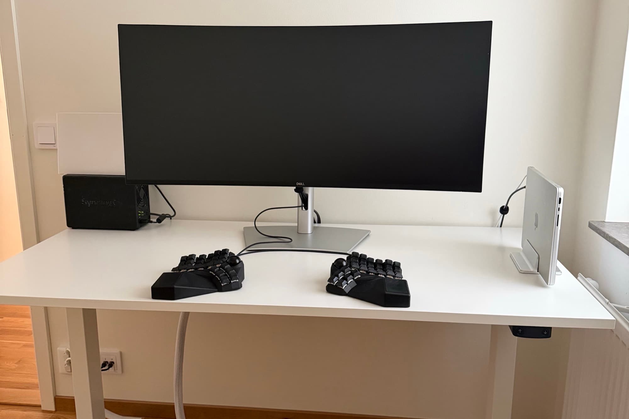 A white sit-stand desk against a white wall: a curved ultrawide monitor on its stand, a split keyboard in two halves either side of the centre, and a laptop closed in a vertical stand to the right.